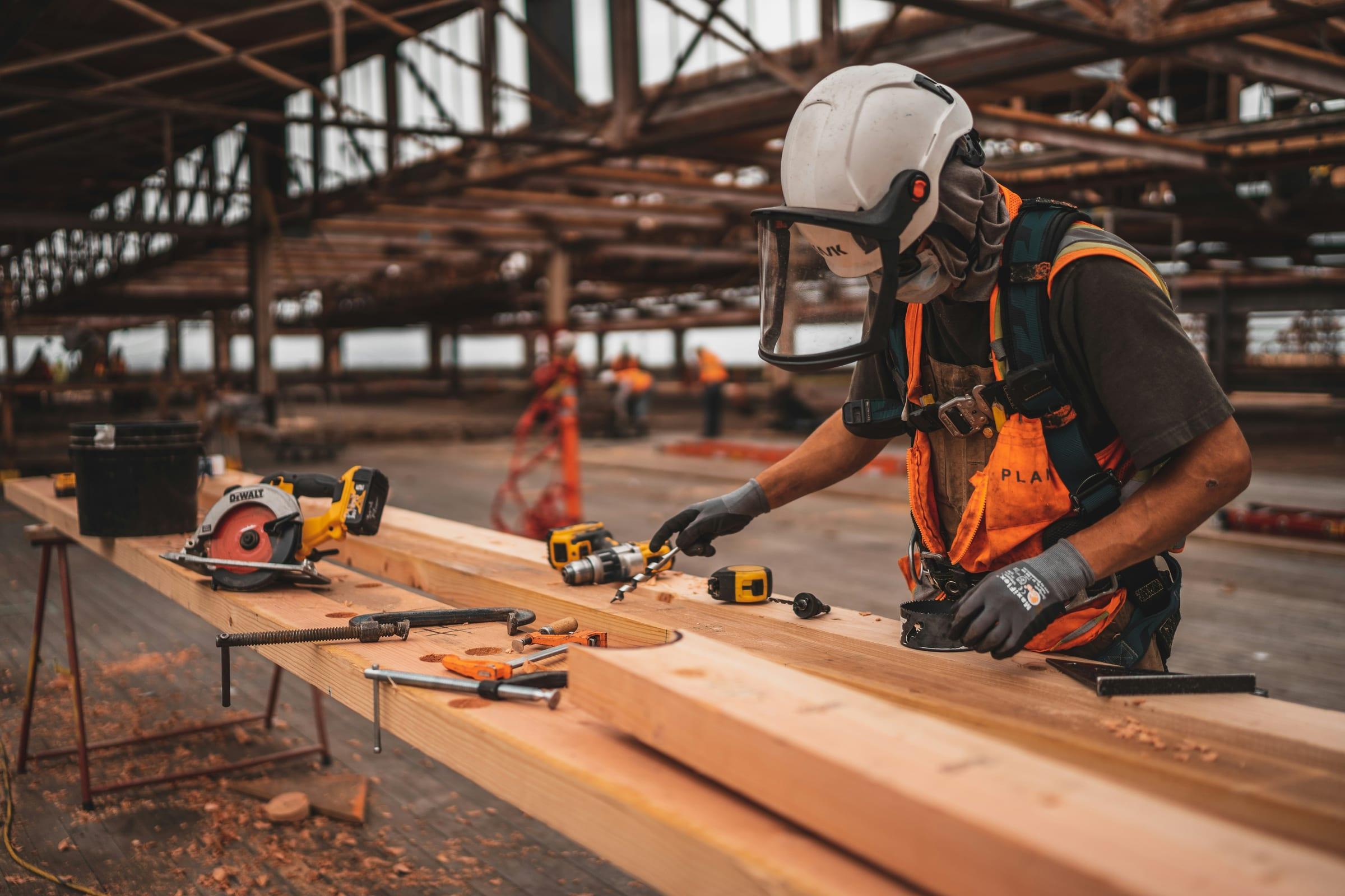 A worker in a hard hat and face shield cutting timber on site.